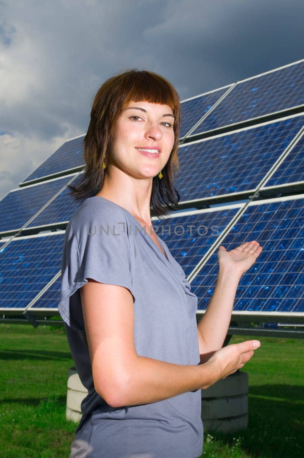Lateral upper body view of a young woman with her hands on a photovoltaic panel standing behind her pointing and smiling friendly into the camera.