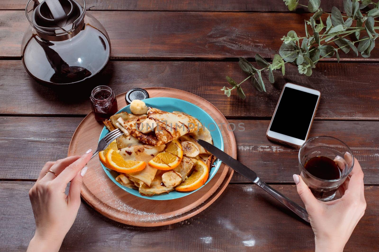 The female hands, phone and pancakes with juice nd female hands. Healthy breakfast. Fresh homemade french crepes made with ffresh fruits on a vintage wooden board on the wooden table. Top view with honey, fruits and coffee. French pancakes concept