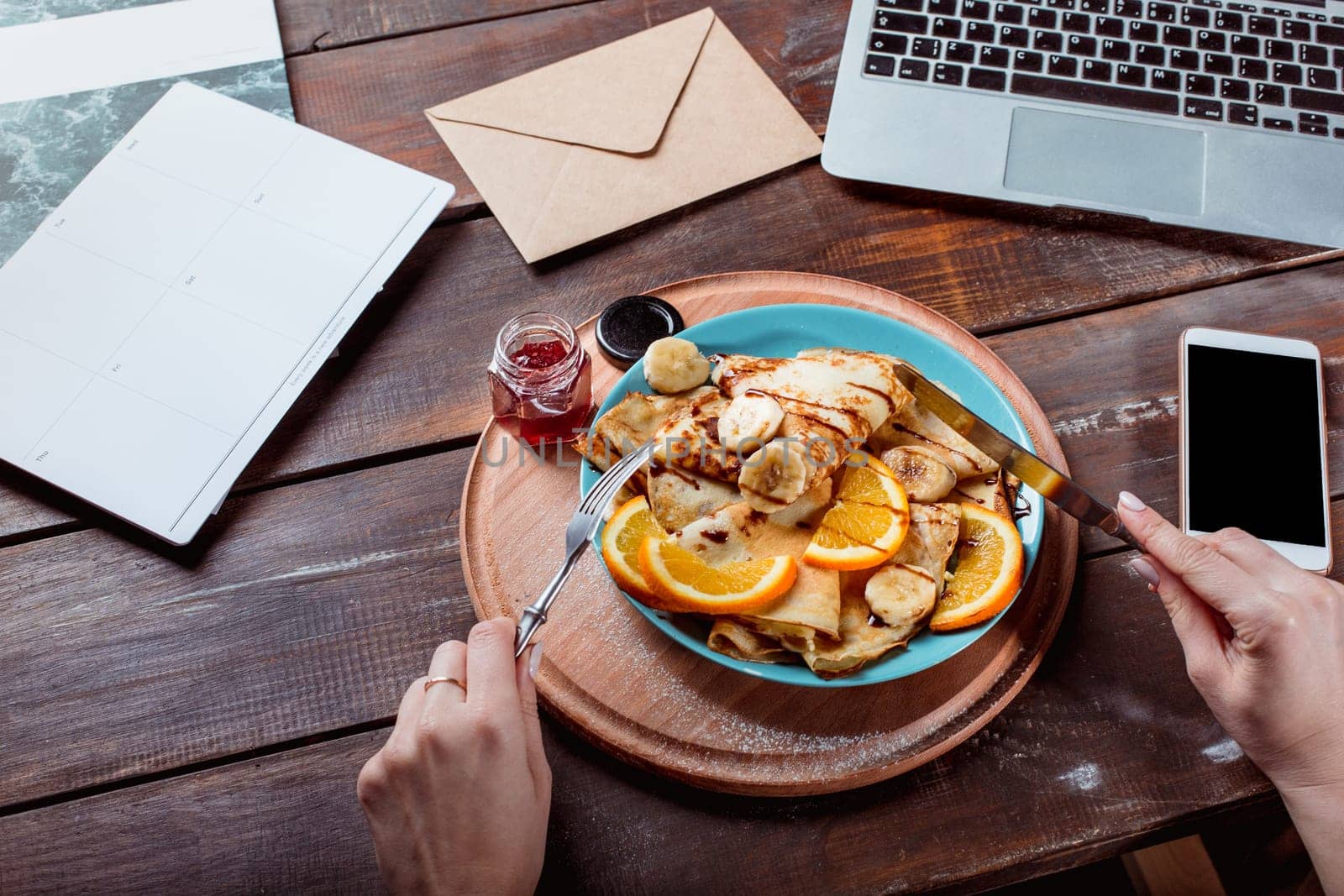 Laptop and pancakes with juice nd female hands. Healthy breakfast. Fresh homemade french crepes made with ffresh fruits on a vintage wooden board on the wooden table. Top view with honey, fruits and coffee. French pancakes concept