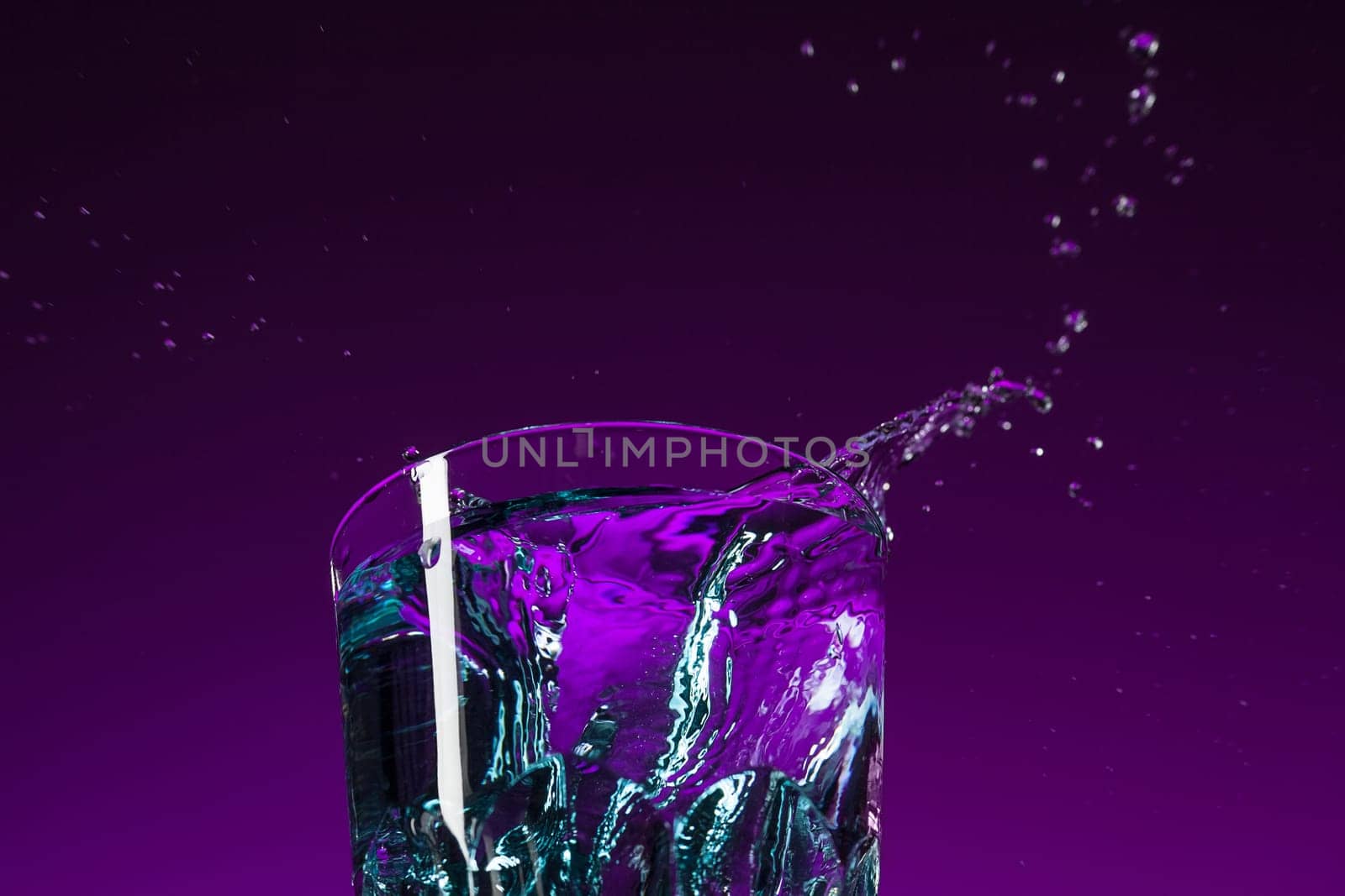 The water splashing in glass on lilac background at studio. Vivid bright colored lighting. Trendy in 2018 Ultra Violet Light Bulb. Art decoration with tone of mystic color