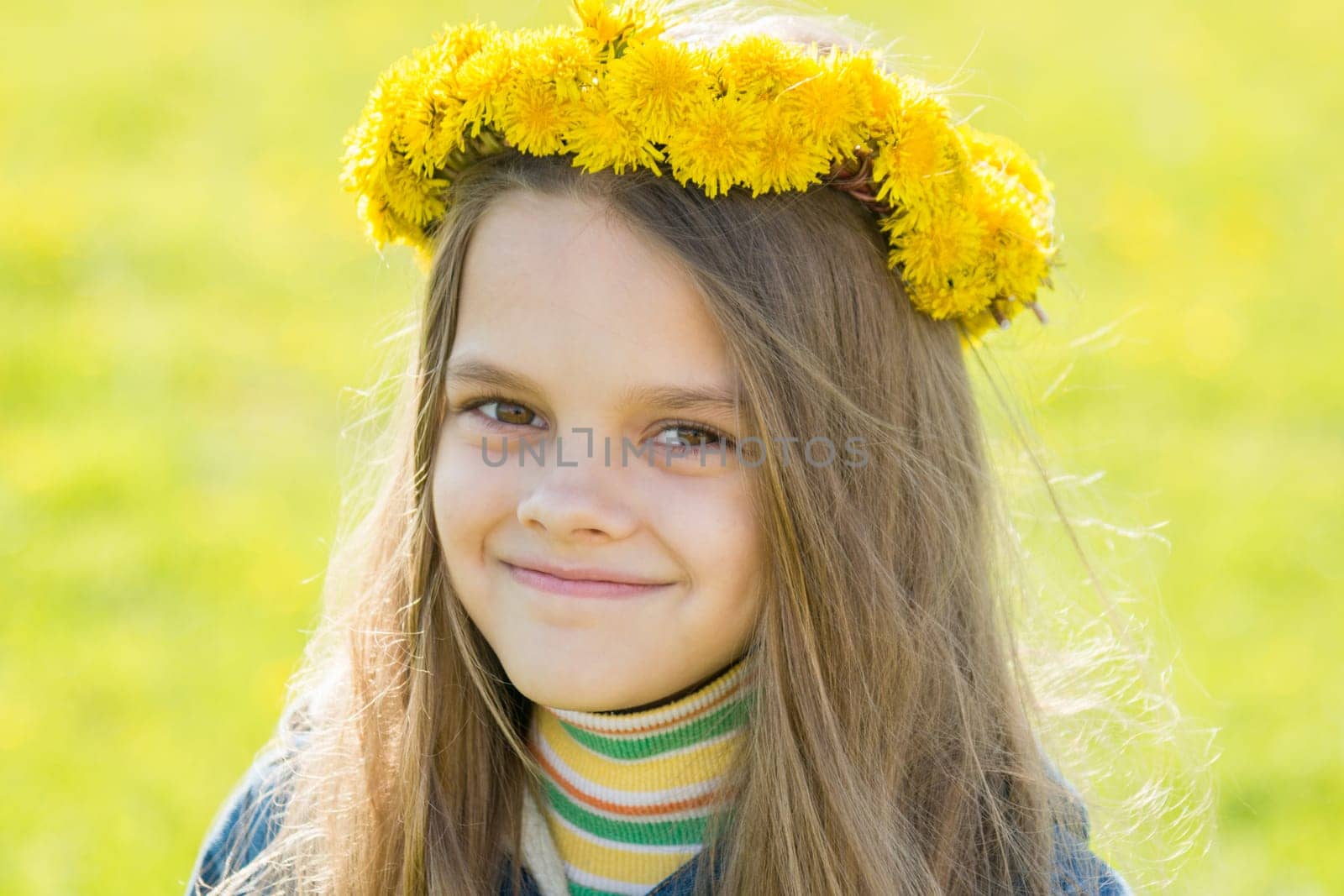 Portrait of a happy eight-year-old girl with a wreath of dandelions on her head, against the background of a spring clearing