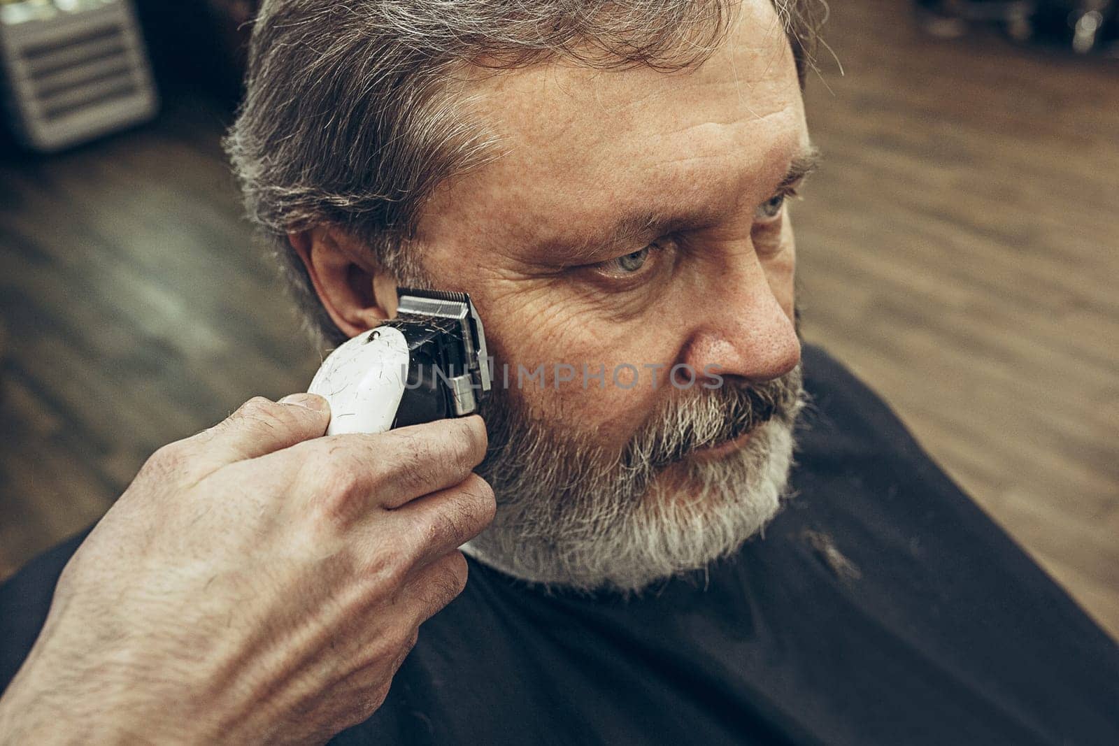 Close-up side view portrait of handsome senior bearded caucasian man getting beard grooming in modern barbershop. Hairdresser serving client, making beard haircut using shaving machine. Barber shop concept.