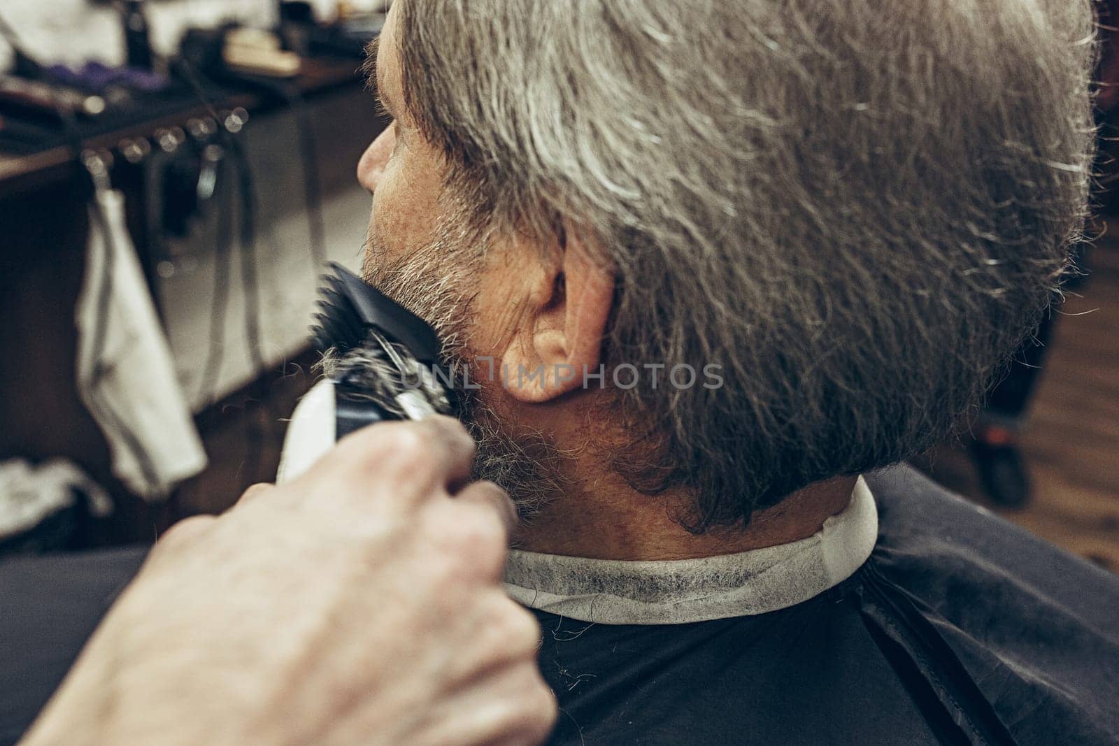Close-up side back view handsome senior bearded caucasian man getting beard grooming in modern barbershop. Hairdresser serving client, making beard haircut using shaving machine. Barber shop concept.