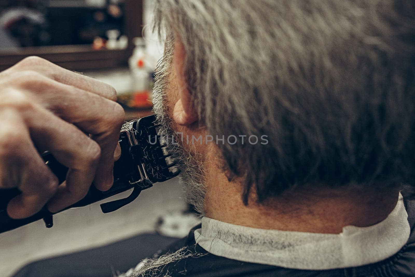 Close-up side back view handsome senior bearded caucasian man getting beard grooming in modern barbershop. Hairdresser serving client, making beard haircut using shaving machine. Barber shop concept.