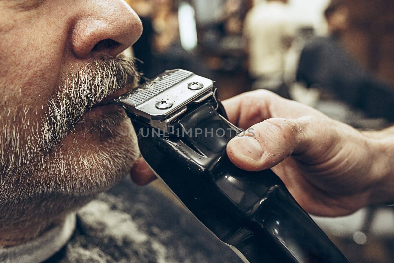 Close-up side view portrait of handsome senior bearded caucasian man getting beard grooming in modern barbershop. Hairdresser serving client, making beard haircut using shaving machine. Barber shop concept.