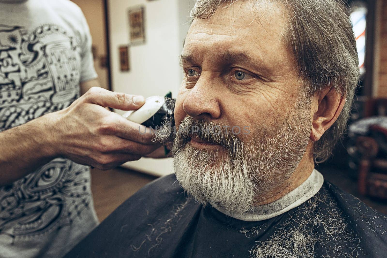 Close-up side view portrait of handsome senior bearded caucasian man getting beard grooming in modern barbershop. Hairdresser serving client, making beard haircut using shaving machine. Barber shop concept.