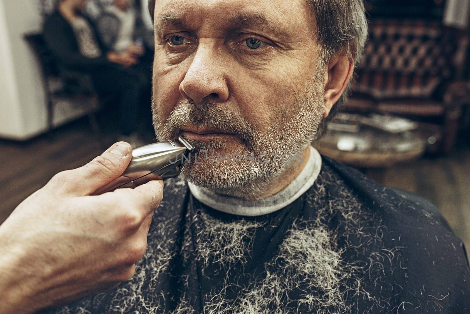 Close-up side view portrait of handsome senior bearded caucasian man getting beard grooming in modern barbershop. Hairdresser serving client, making beard haircut using shaving machine. Barber shop concept.