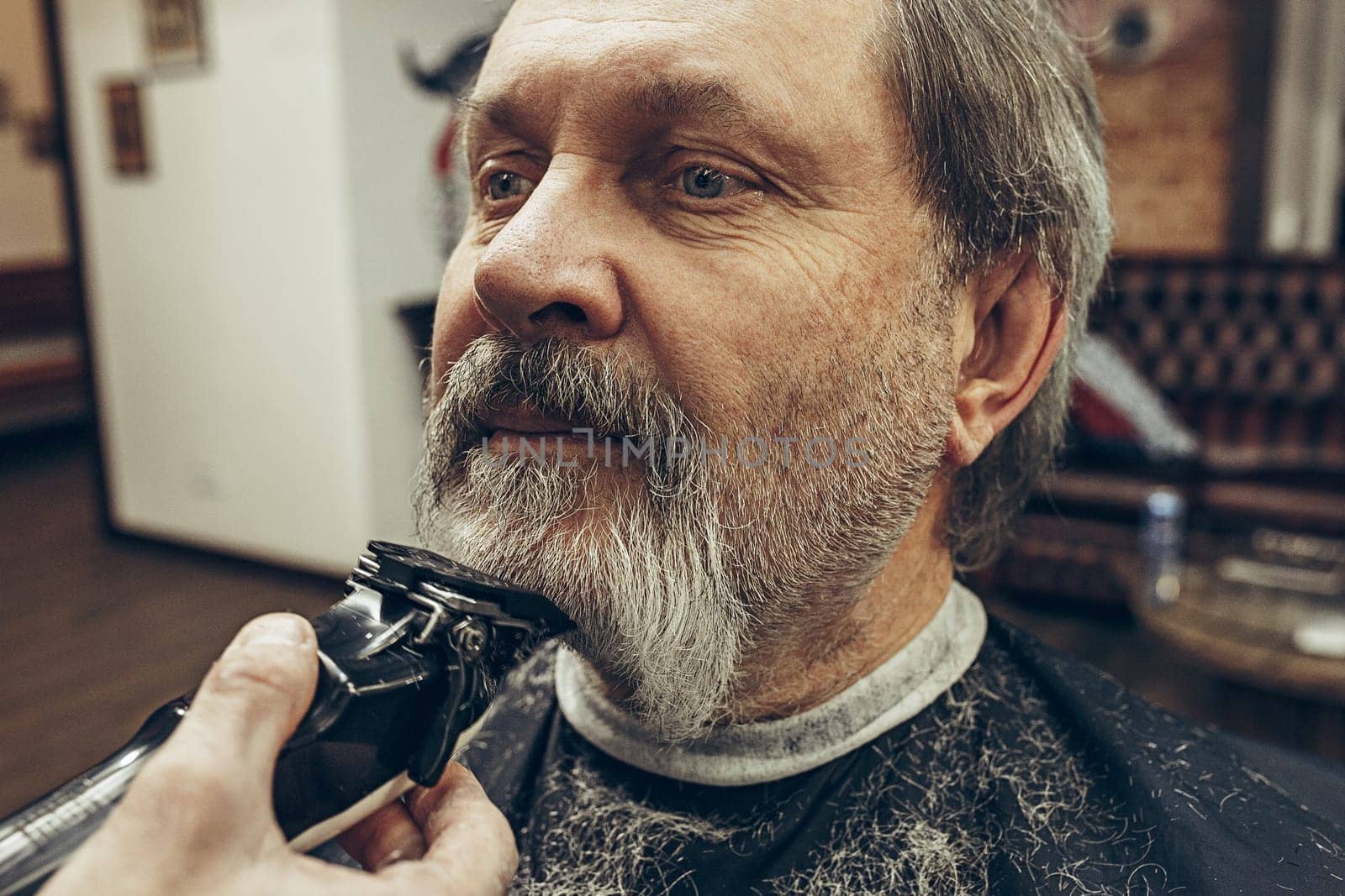 Close-up side view portrait of handsome senior bearded caucasian man getting beard grooming in modern barbershop. Hairdresser serving client, making beard haircut using shaving machine. Barber shop concept.