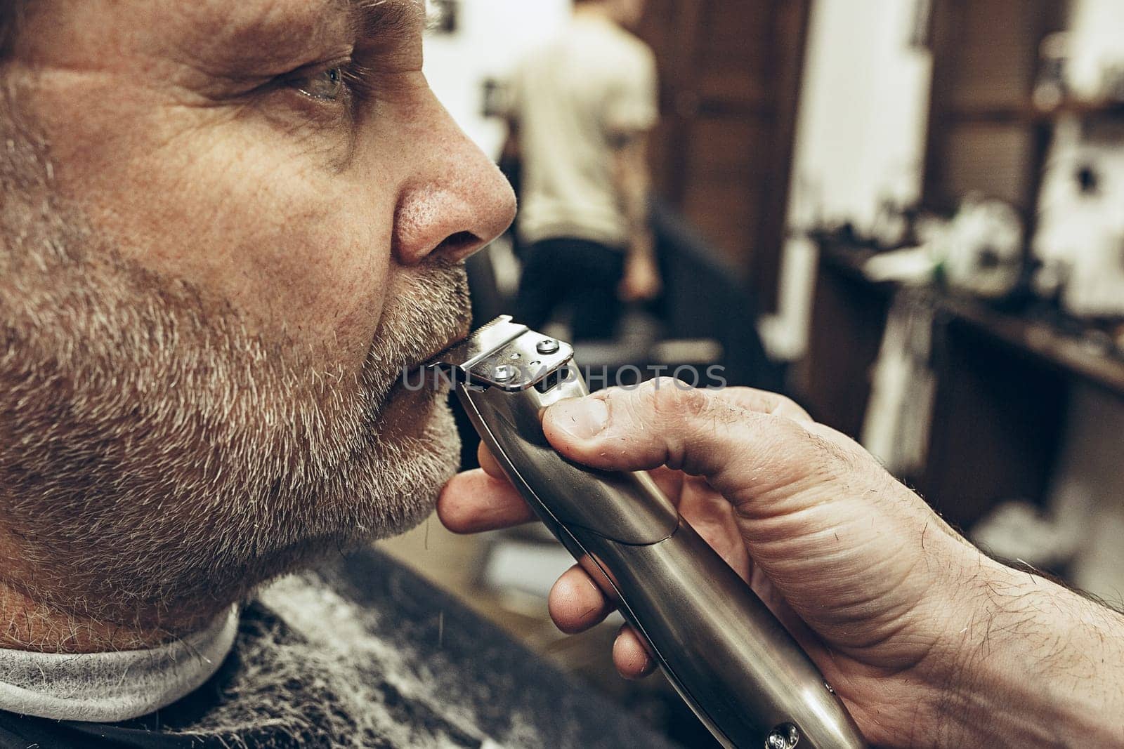 Close-up side profile portrait of handsome senior bearded caucasian man getting beard grooming in modern barbershop. Hairdresser serving client, making beard haircut using shaving machine. Barber shop concept.