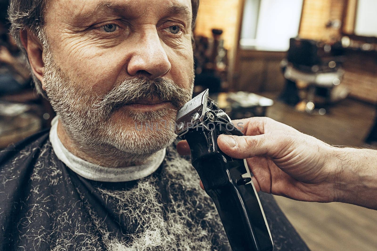 Close-up side view portrait of handsome senior bearded caucasian man getting beard grooming in modern barbershop. Hairdresser serving client, making beard haircut using shaving machine. Barber shop concept.