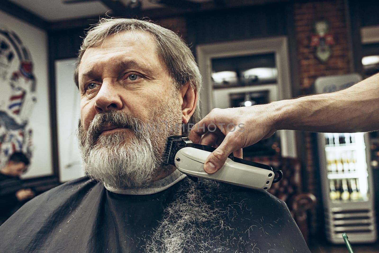 Close-up side view portrait of handsome senior bearded caucasian man getting beard grooming in modern barbershop. Hairdresser serving client, making beard haircut using shaving machine. Barber shop concept.