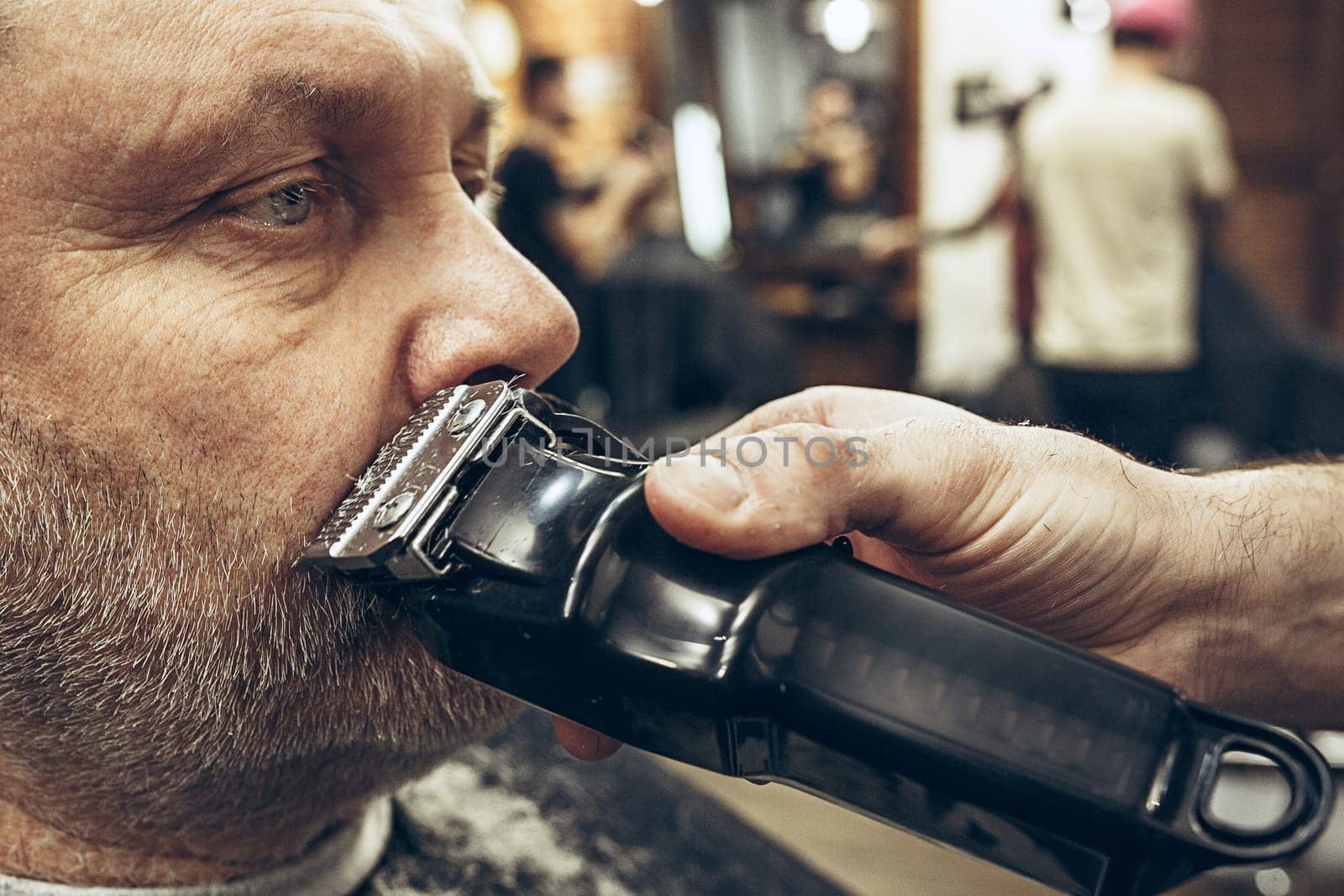 Close-up side view portrait of handsome senior bearded caucasian man getting beard grooming in modern barbershop. Hairdresser serving client, making beard haircut using shaving machine. Barber shop concept.