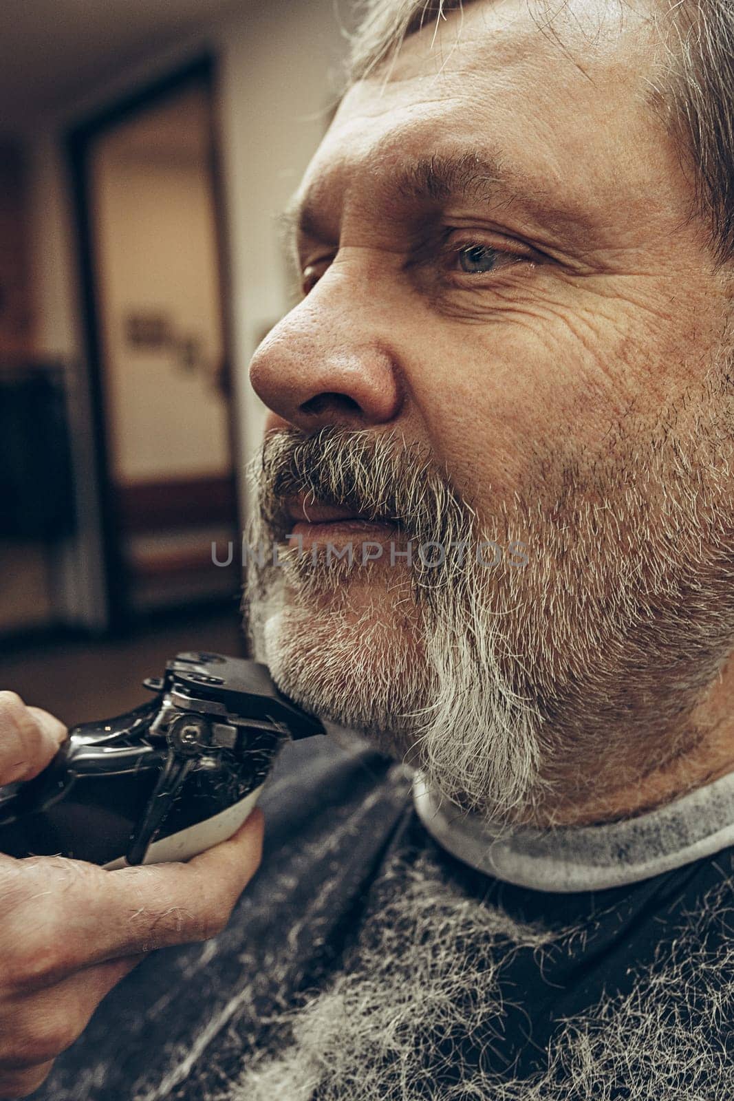 Close-up side view portrait of handsome senior bearded caucasian man getting beard grooming in modern barbershop. Hairdresser serving client, making beard haircut using shaving machine. Barber shop concept.