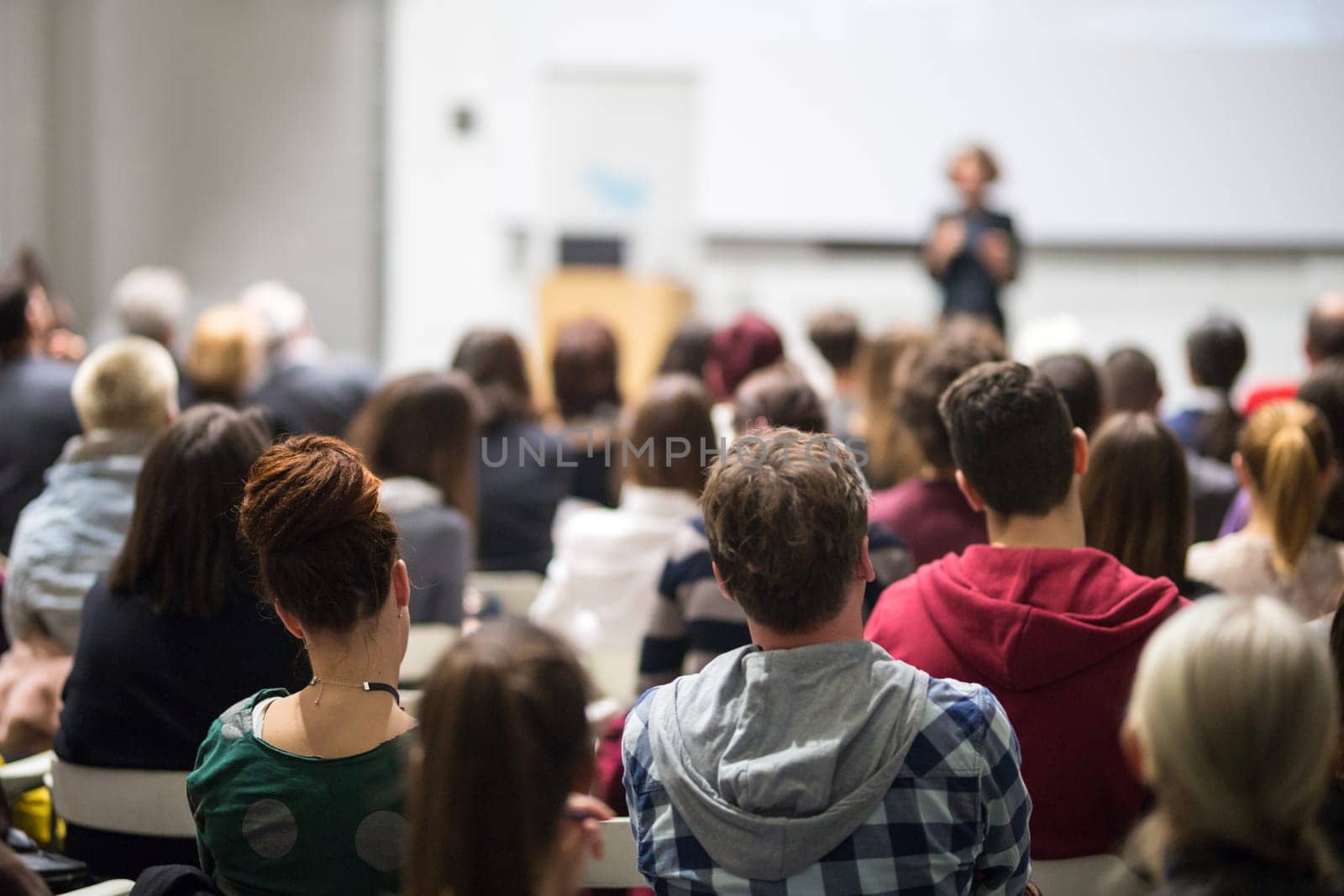 Female speaker giving presentation in lecture hall at university workshop. Audience in conference hall. Rear view of unrecognized participant in audience. Scientific conference event.