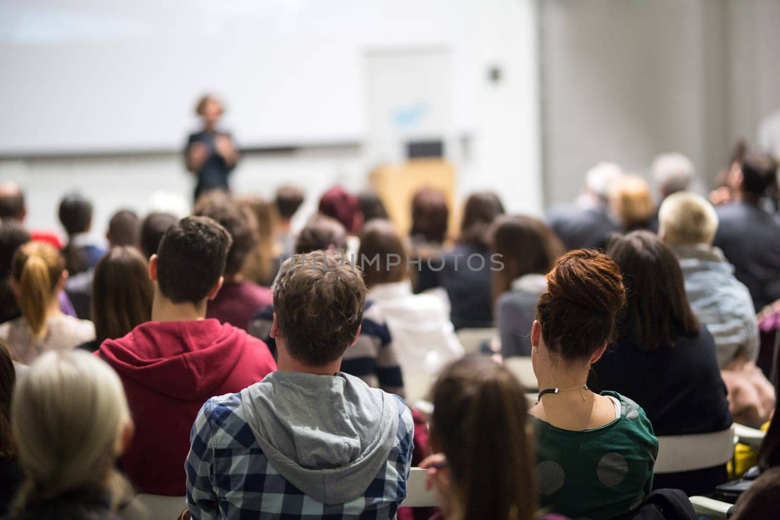 Female speaker giving presentation in lecture hall at university workshop. Audience in conference hall. Rear view of unrecognized participant in audience. Scientific conference event.