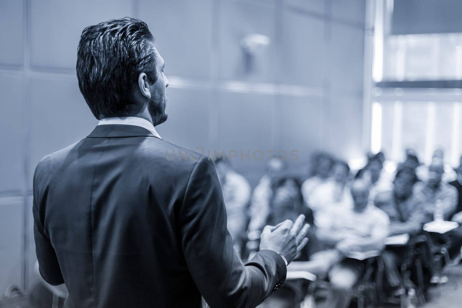 Rear view of speaker giving a talk at business meeting. Audience in the conference hall. Business and Entrepreneurship concept. Black and white blue toned image.