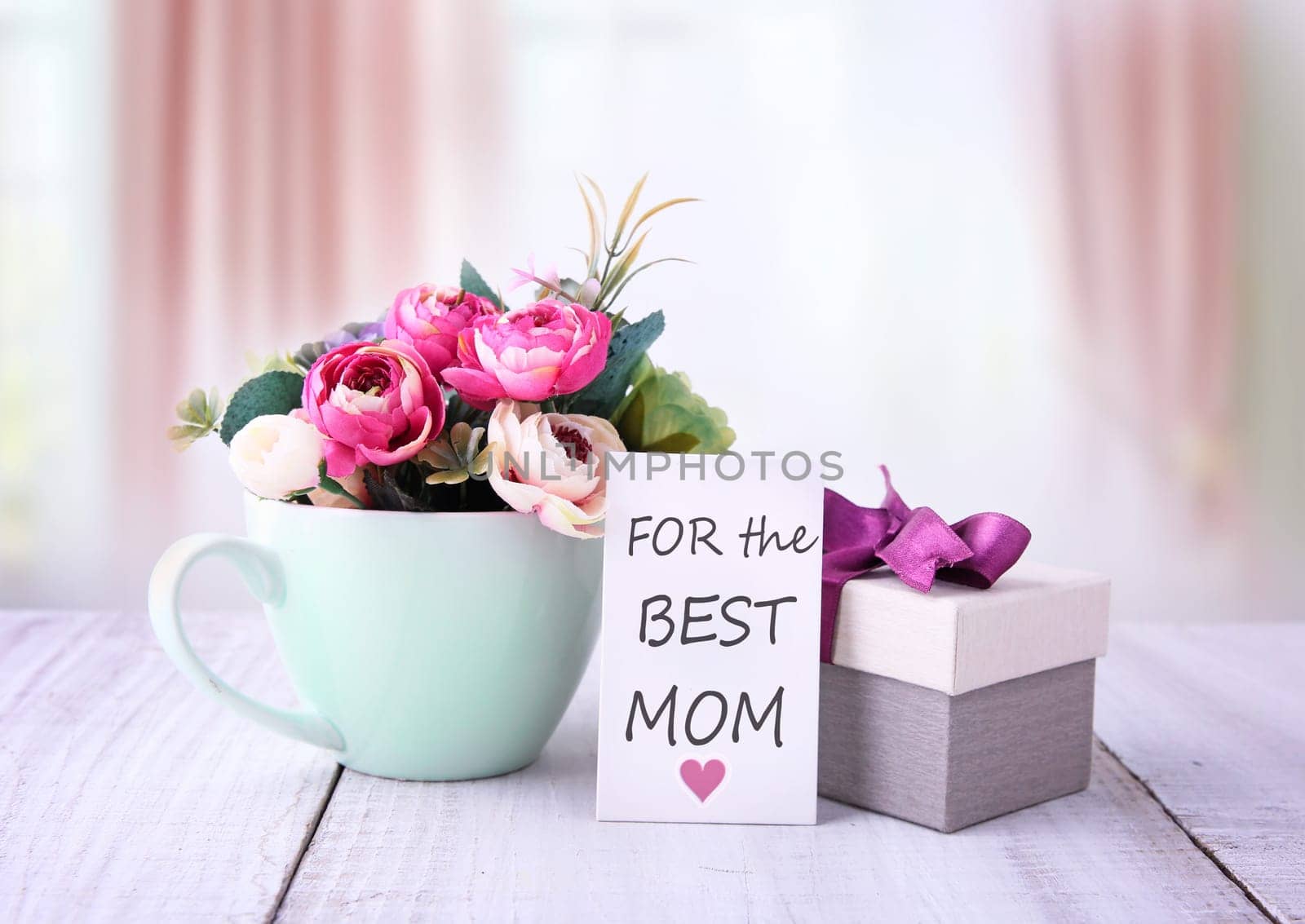 Mother's day greeting card.Birthday gift box with flowers on wooden table. Happy mothers day gifts with red white roses in a cup, and gift tag with text „For the best mom“, presented on wooden table with bright backround
