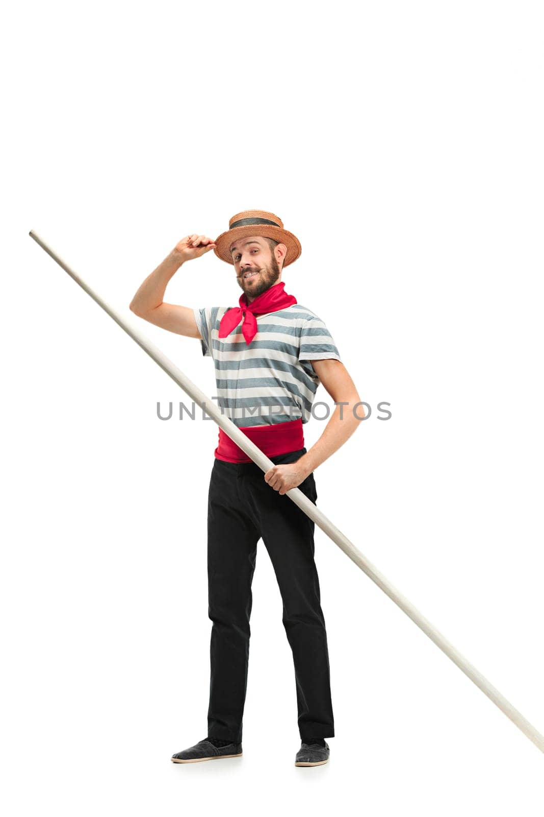 Caucasian funny bearded man in traditional gondolier costume and hat with paddle standing and posing at studio isolated on white background. Smiling young man. Human emotions, facial expression concept