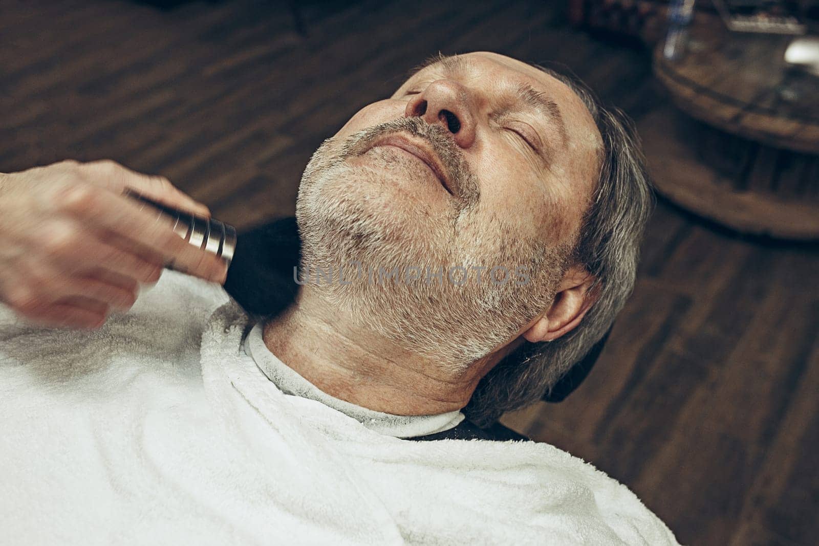 Close-up side top view handsome senior bearded caucasian man getting beard grooming in modern barbershop. Hairdresser serving client, making beard haircut using straight razor. Barber shop concept.