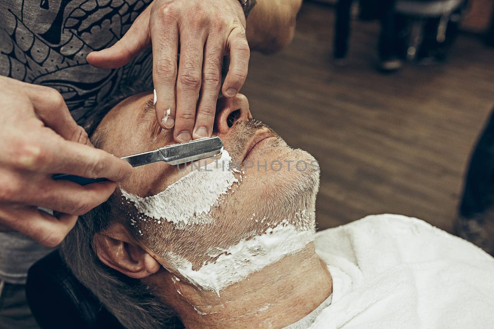 Close-up side top view handsome senior bearded caucasian man getting beard grooming in modern barbershop. Hairdresser serving client, making beard haircut using straight razor. Barber shop concept.