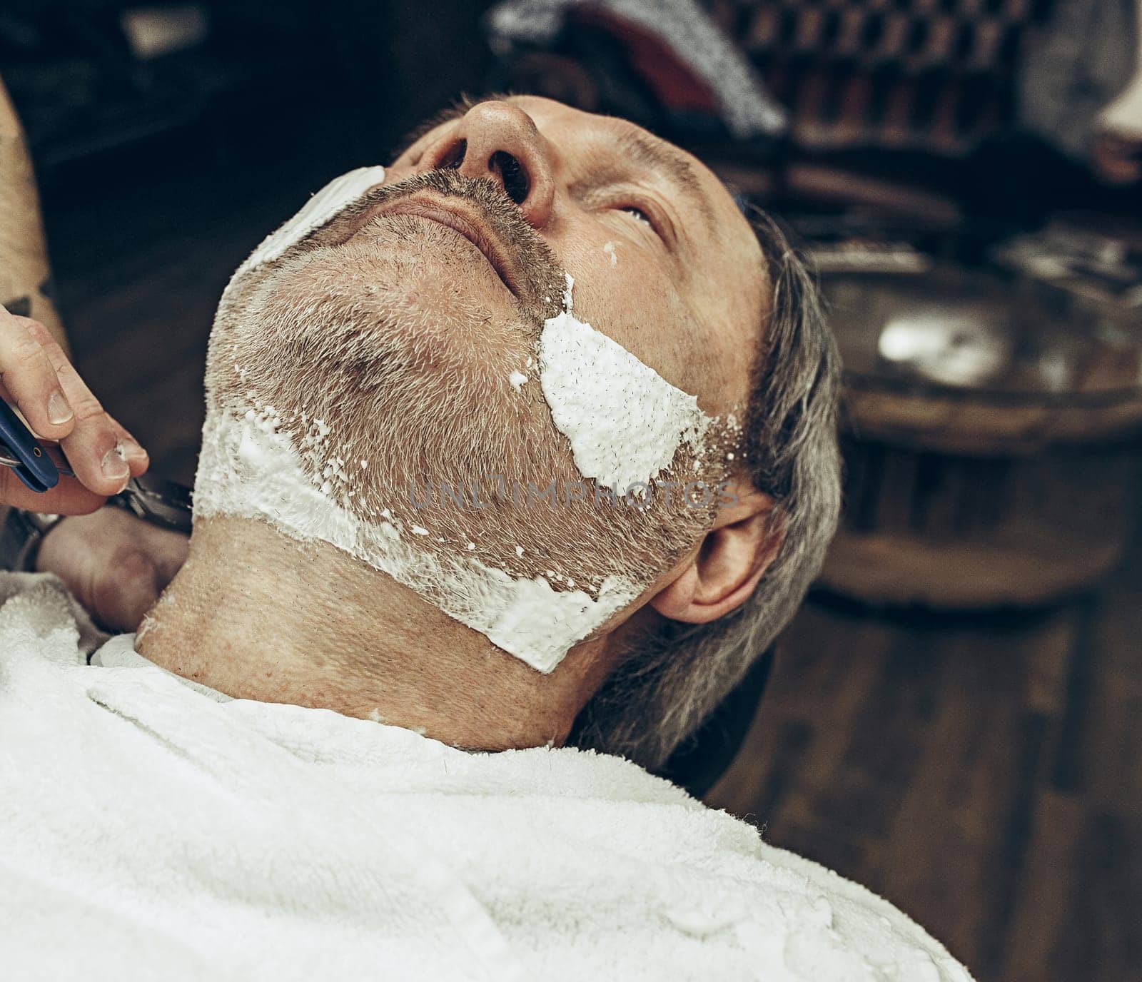 Close-up side top view handsome senior bearded caucasian man getting beard grooming in modern barbershop. Hairdresser serving client, making beard haircut using straight razor. Barber shop concept.
