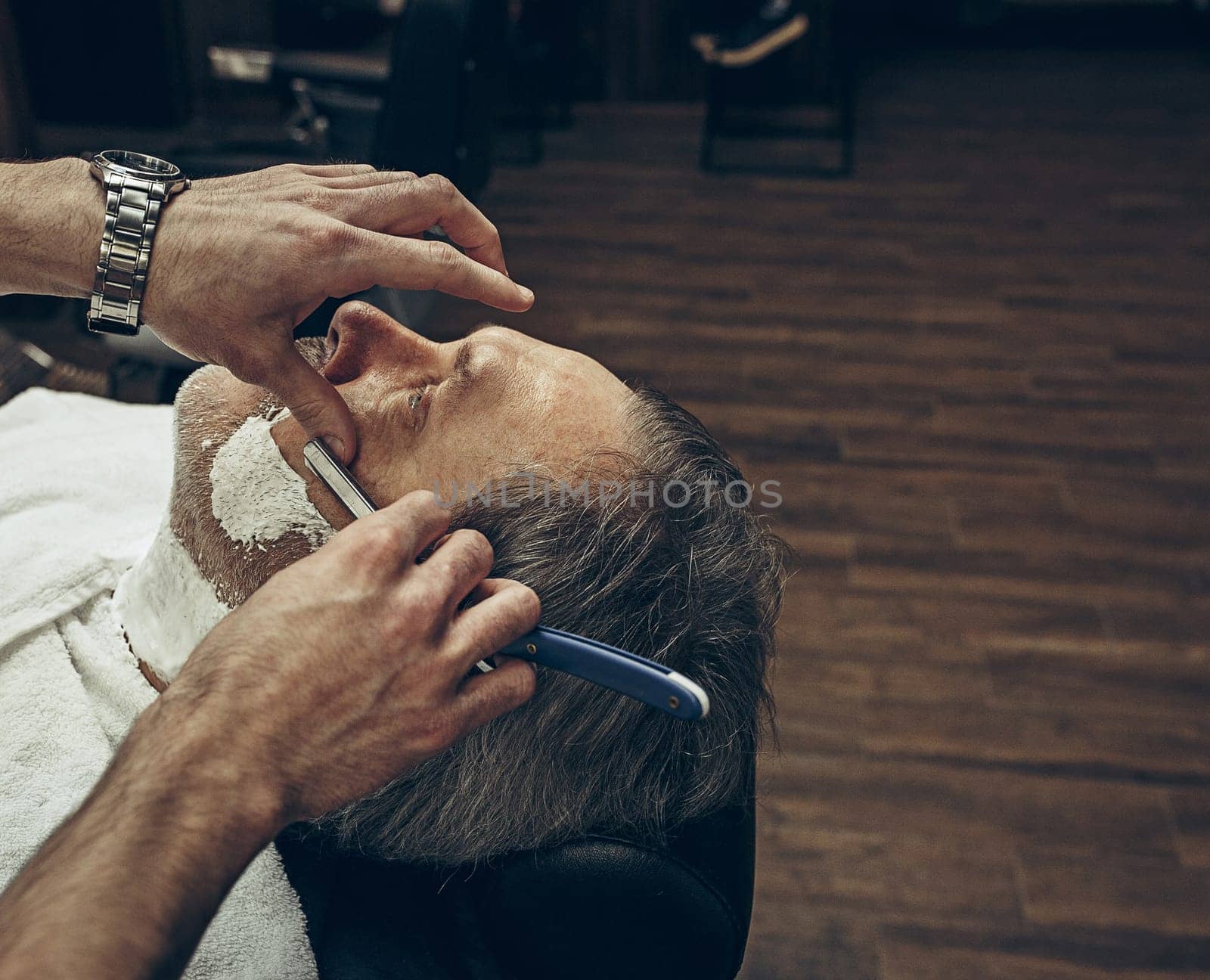 Close-up side top view handsome senior bearded caucasian man getting beard grooming in modern barbershop. Hairdresser serving client, making beard haircut using straight razor. Barber shop concept.