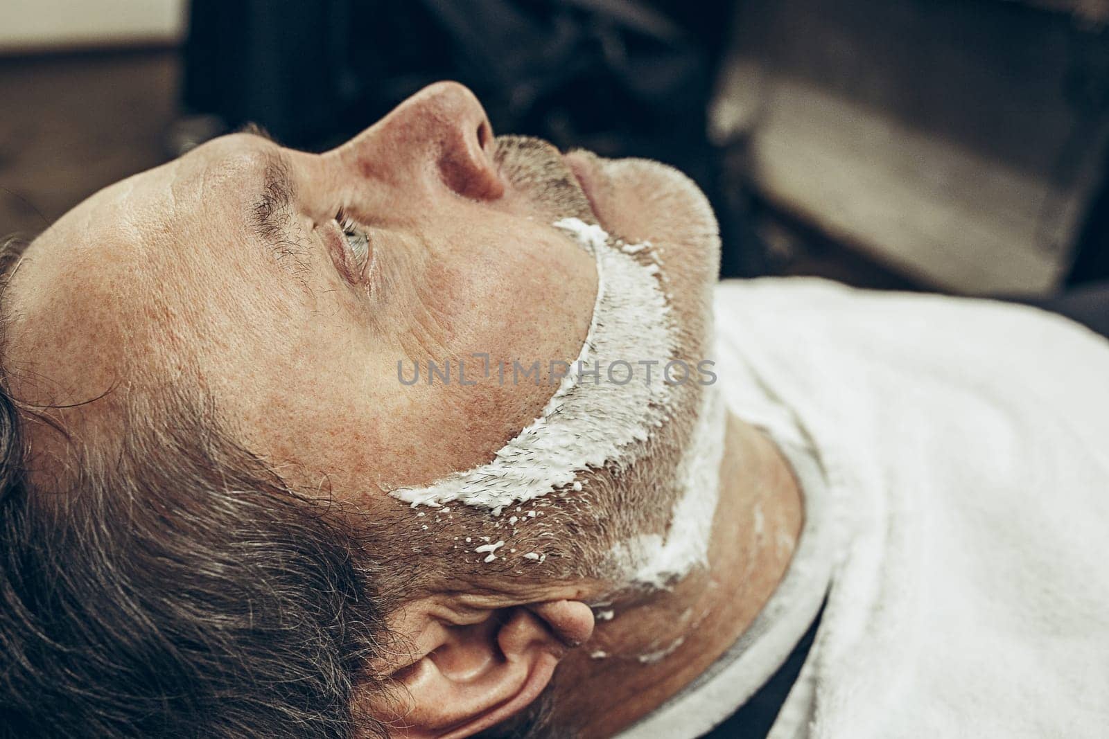 Close-up side top view handsome senior bearded caucasian man getting beard grooming in modern barbershop. Hairdresser serving client, making beard haircut using straight razor. Barber shop concept.