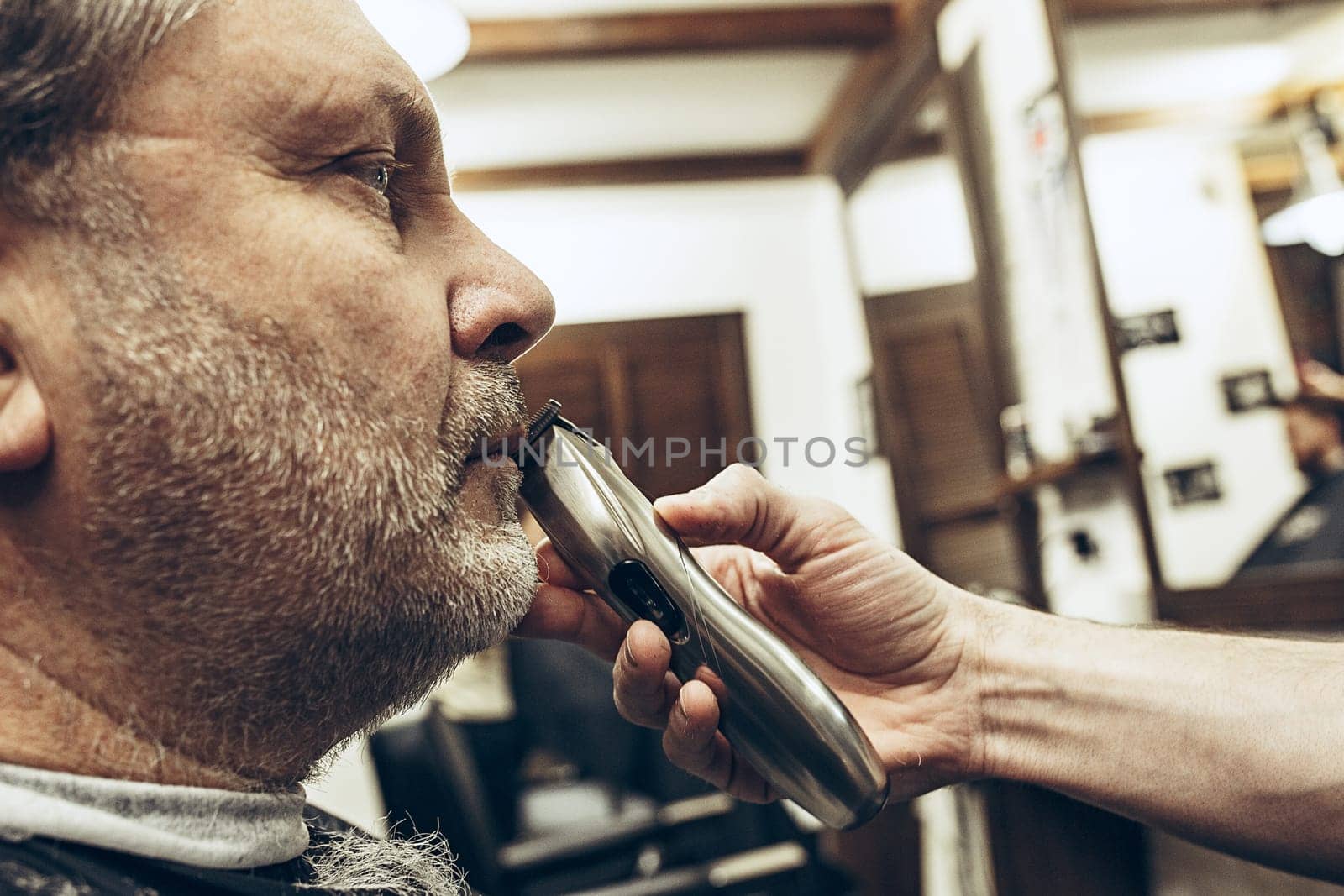 Close-up side profile portrait of handsome senior bearded caucasian man getting beard grooming in modern barbershop. Hairdresser serving client, making beard haircut using shaving machine. Barber shop concept.