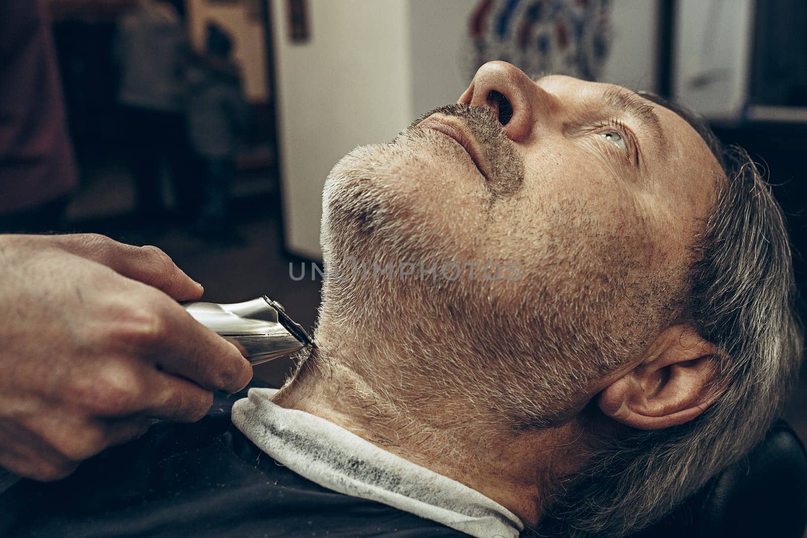 Close-up side profile portrait of handsome senior bearded caucasian man getting beard grooming in modern barbershop. Hairdresser serving client, making beard haircut using shaving machine. Barber shop concept.