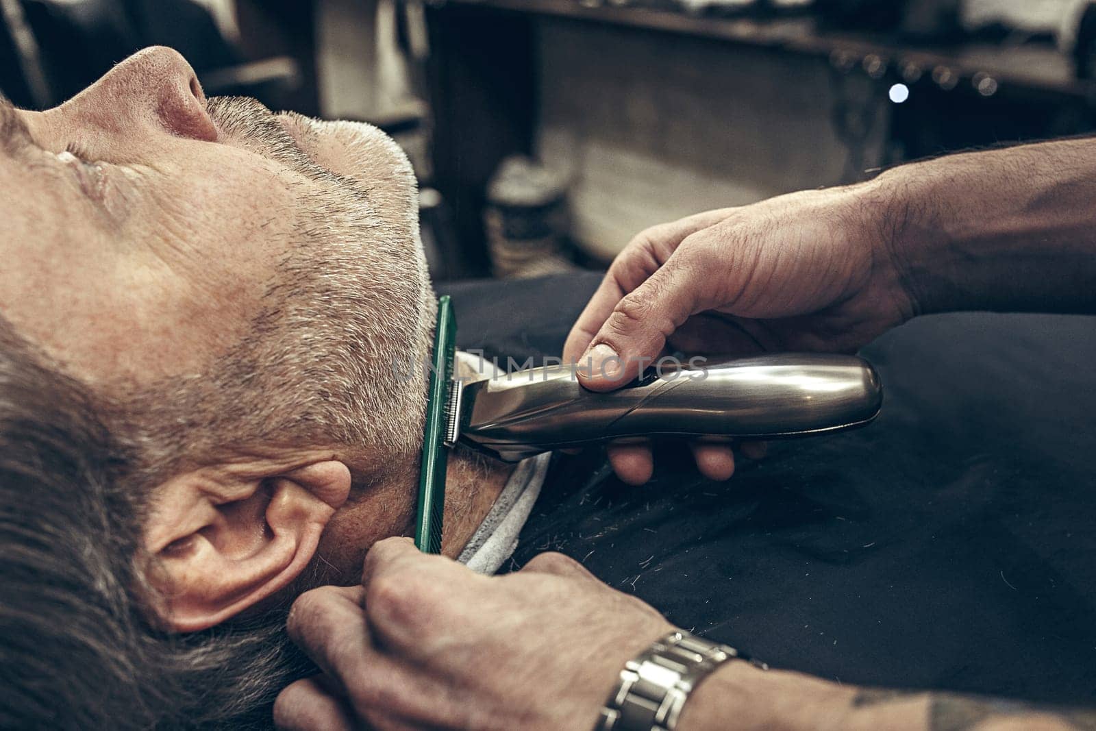 Close-up side profile portrait of handsome senior bearded caucasian man getting beard grooming in modern barbershop. Hairdresser serving client, making beard haircut using shaving machine. Barber shop concept.