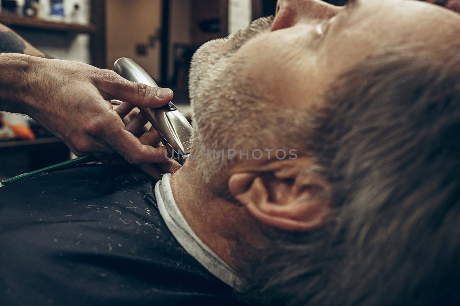 Close-up side back view handsome senior bearded caucasian man getting beard grooming in modern barbershop. Hairdresser serving client, making beard haircut using shaving machine. Barber shop concept.