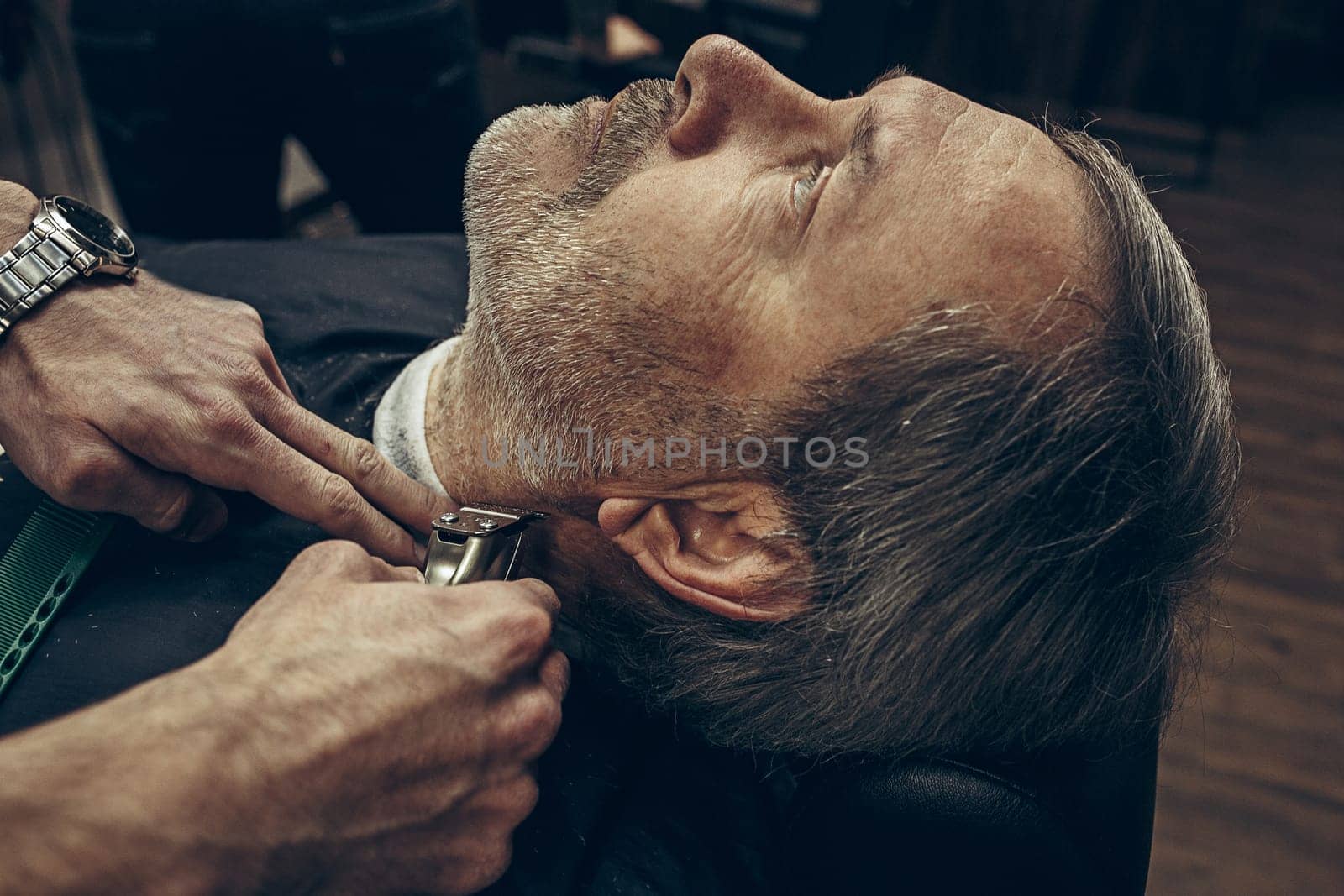 Close-up side back view handsome senior bearded caucasian man getting beard grooming in modern barbershop. Hairdresser serving client, making beard haircut using shaving machine. Barber shop concept.