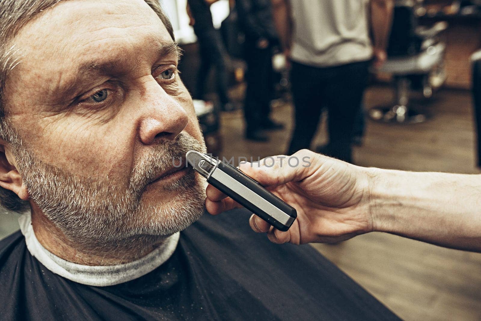 Close-up side view portrait of handsome senior bearded caucasian man getting beard grooming in modern barbershop. Hairdresser serving client, making beard haircut using shaving machine. Barber shop concept.