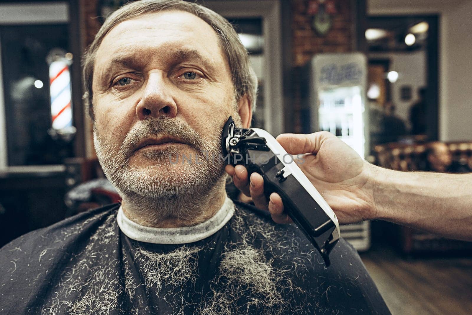 Close-up side view portrait of handsome senior bearded caucasian man getting beard grooming in modern barbershop. Hairdresser serving client, making beard haircut using shaving machine. Barber shop concept.