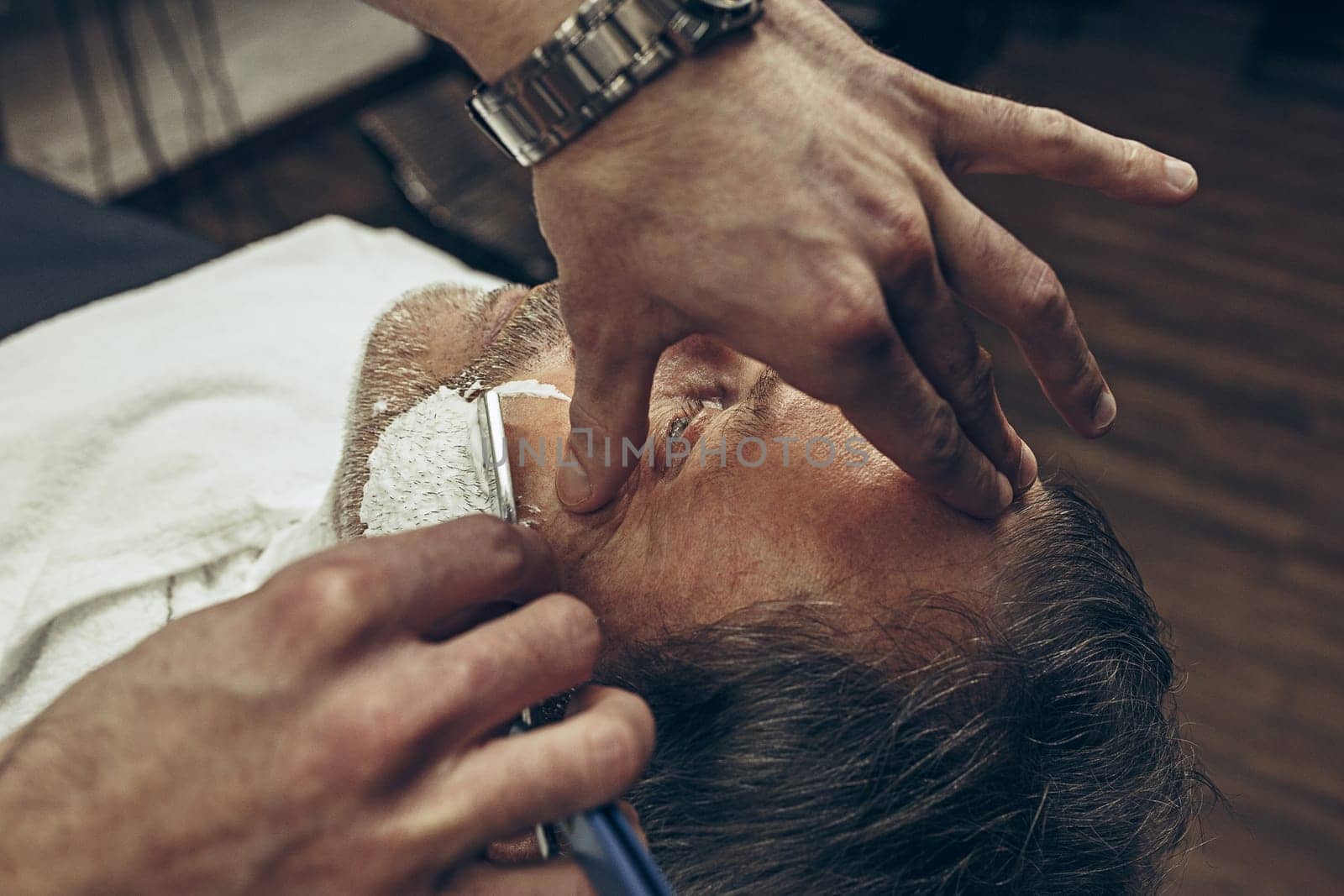 Close-up side top view handsome senior bearded caucasian man getting beard grooming in modern barbershop. Hairdresser serving client, making beard haircut using straight razor. Barber shop concept.