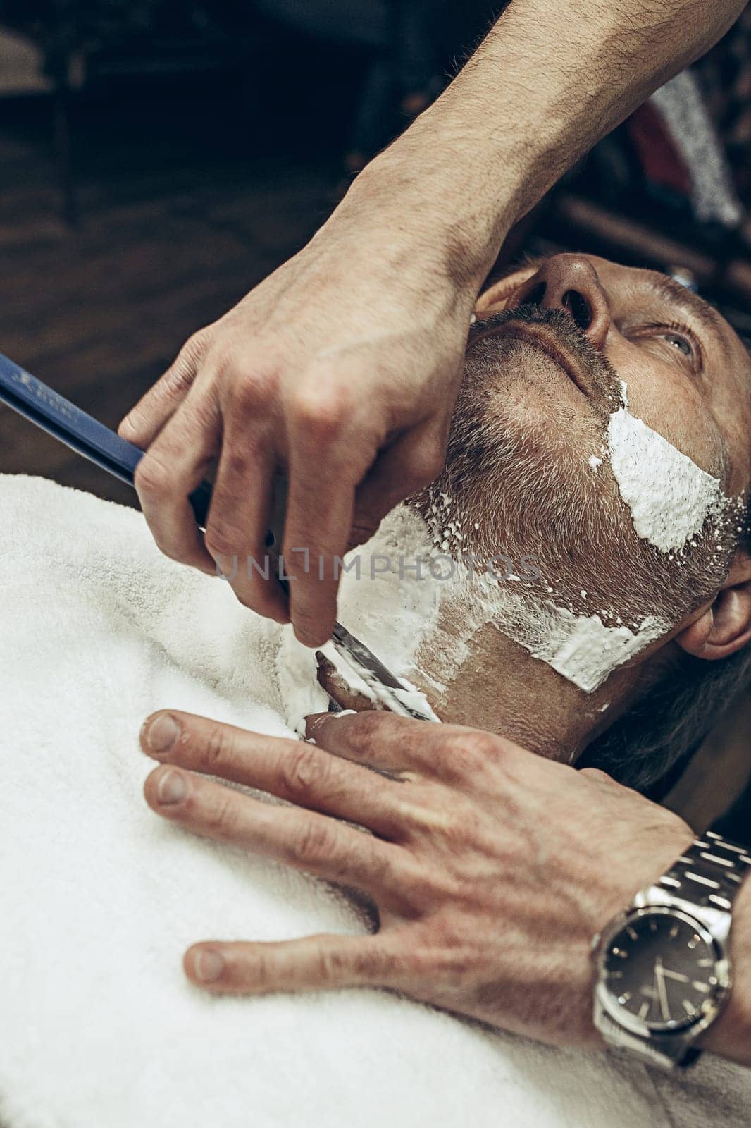 Close-up side top view handsome senior bearded caucasian man getting beard grooming in modern barbershop. Hairdresser serving client, making beard haircut using straight razor. Barber shop concept.