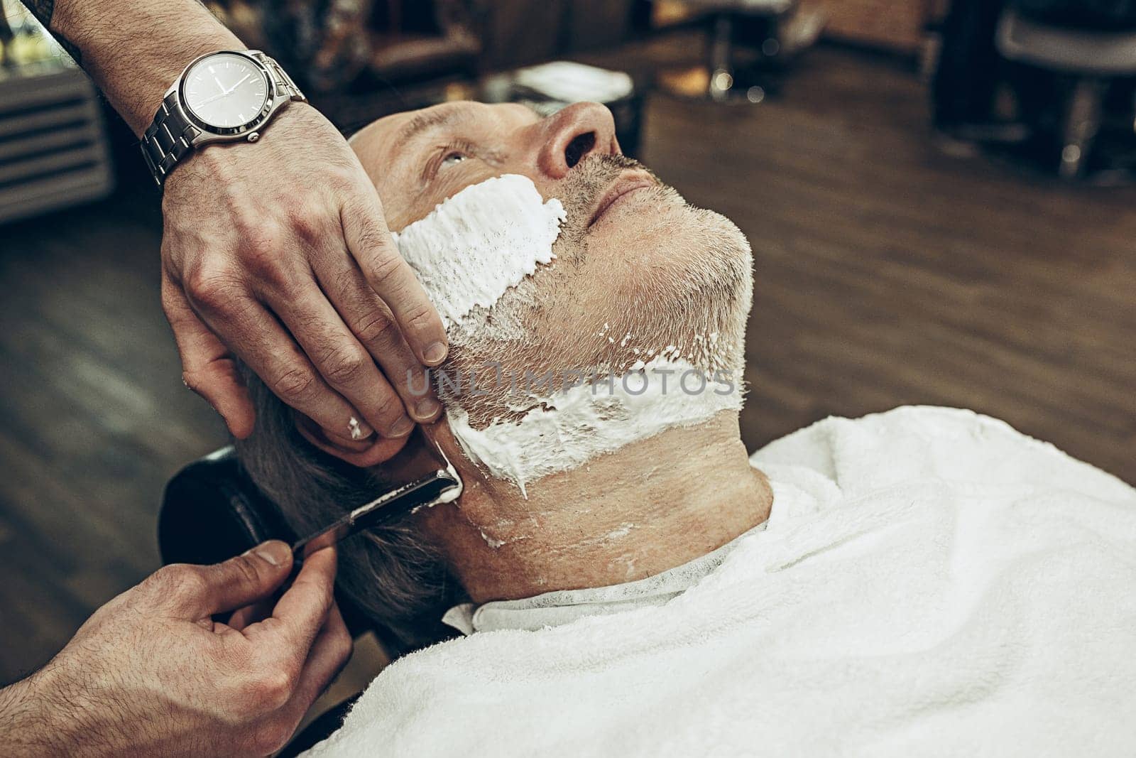 Close-up side top view handsome senior bearded caucasian man getting beard grooming in modern barbershop. Hairdresser serving client, making beard haircut using straight razor. Barber shop concept.