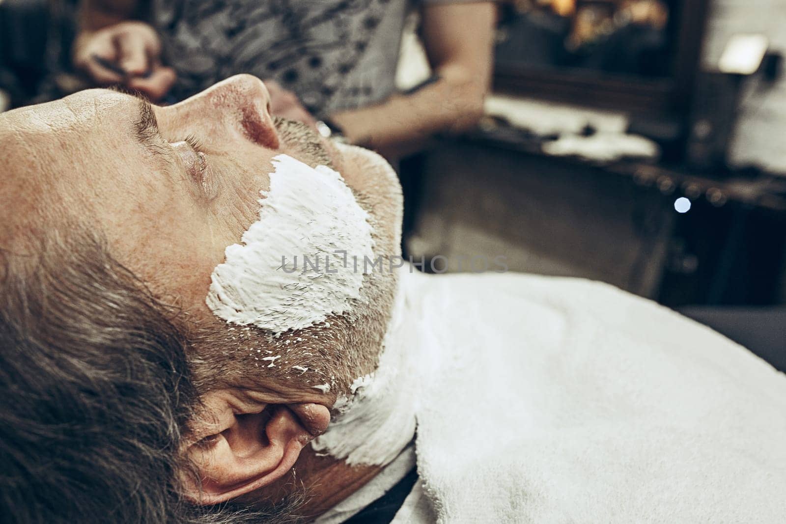Close-up side view portrait of handsome senior bearded caucasian man getting beard grooming in modern barbershop. Hairdresser serving client, making beard haircut using straight razor. Barber shop concept.