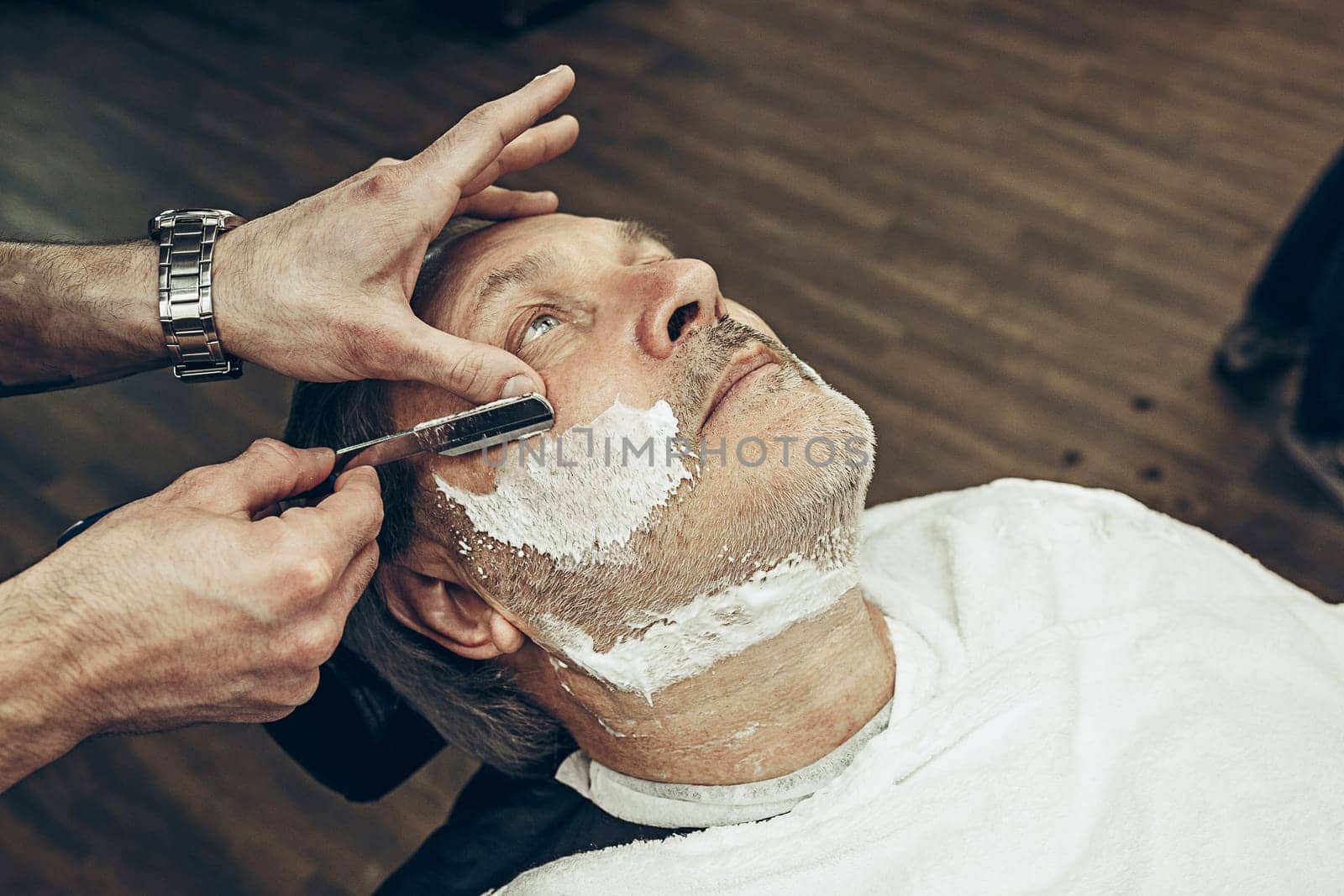 Close-up side top view handsome senior bearded caucasian man getting beard grooming in modern barbershop. Hairdresser serving client, making beard haircut using straight razor. Barber shop concept.