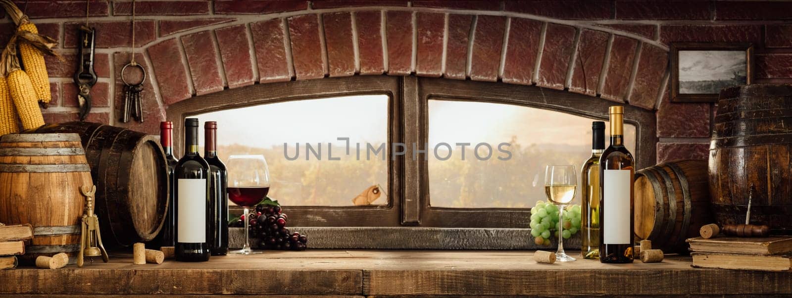 Wine glasses, bottles and barrels in a rustic countryside wine cellar  panoramic window view of lush vineyards at sunset: traditional winemaking concept