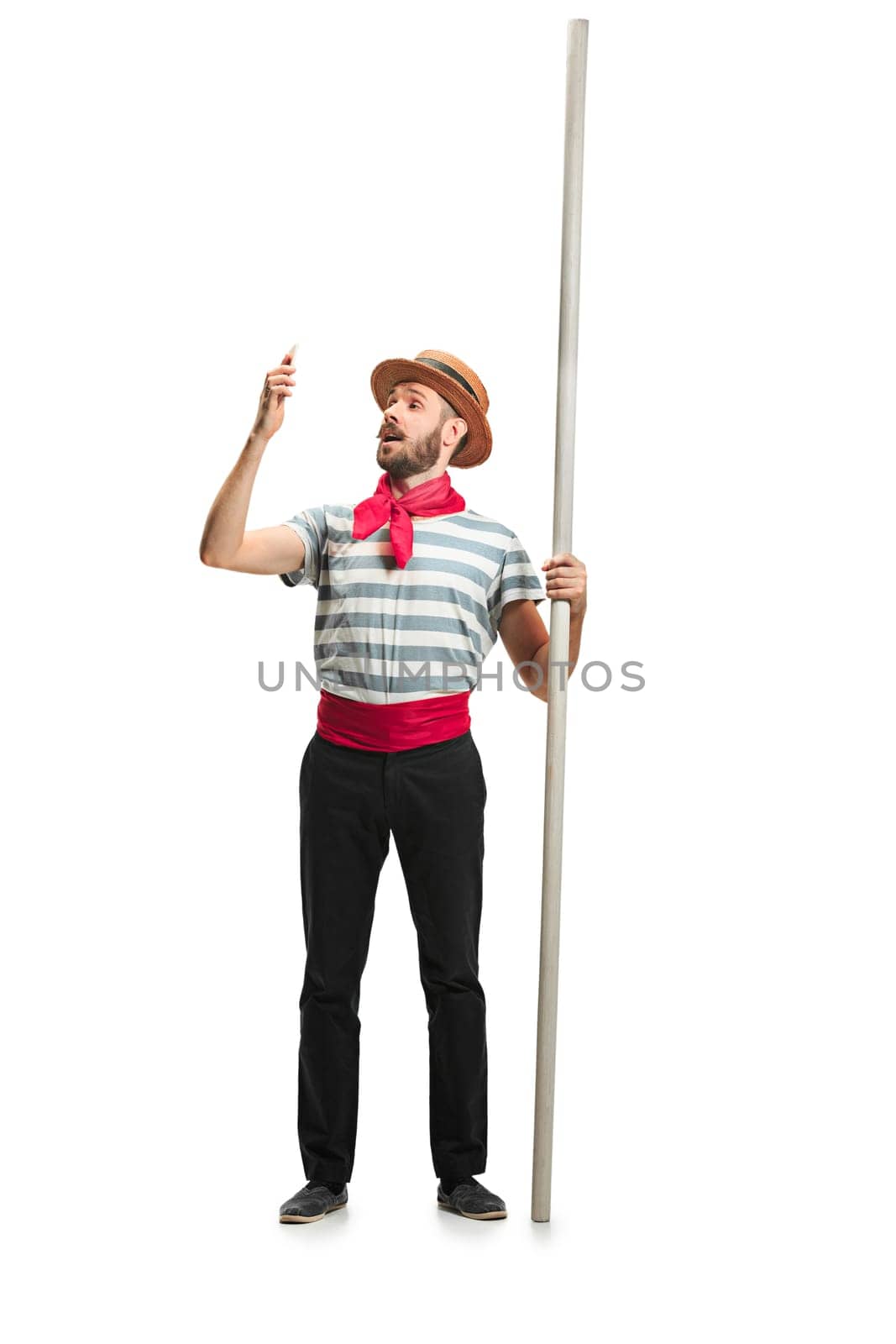 Caucasian funny bearded man in traditional gondolier costume and hat with paddle standing with mobile phone at studio isolated on white background. young man. Human emotions, facial expression concept