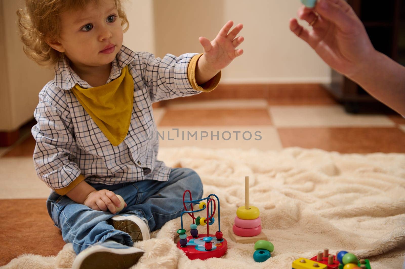 A small child explores colorful educational toys on a blanket, engaging in a happy and focused activity, while a guiding hand supports learning through play.
