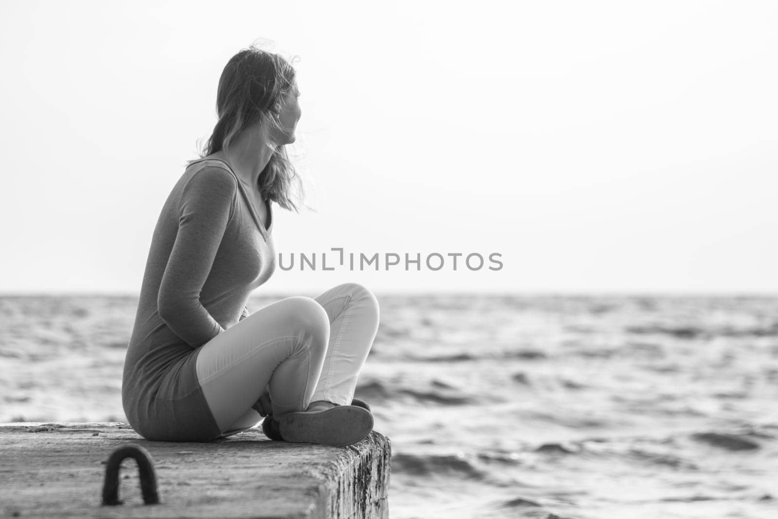 A young girl sits on a pier and looks out into the distance at sea