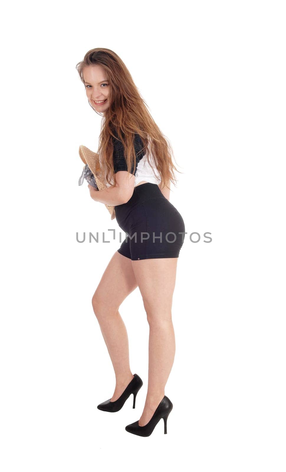 A lovely young woman standing in profile and black shorts with a .hat holding to her chest, isolated for white background.