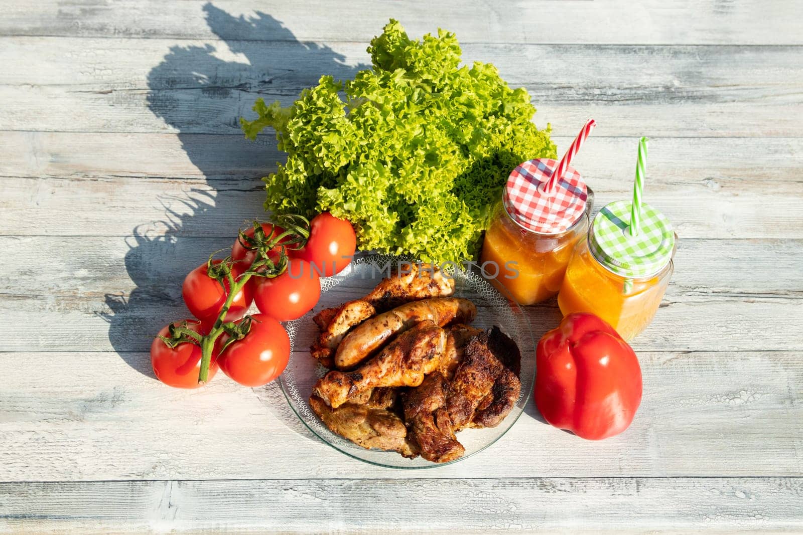 Top view on plate of fresh grilled meat surrounded by fresh salad, tomatoes, paprika and two glasses of freshly squeezed orange juice. presented outdoor on wooden table.