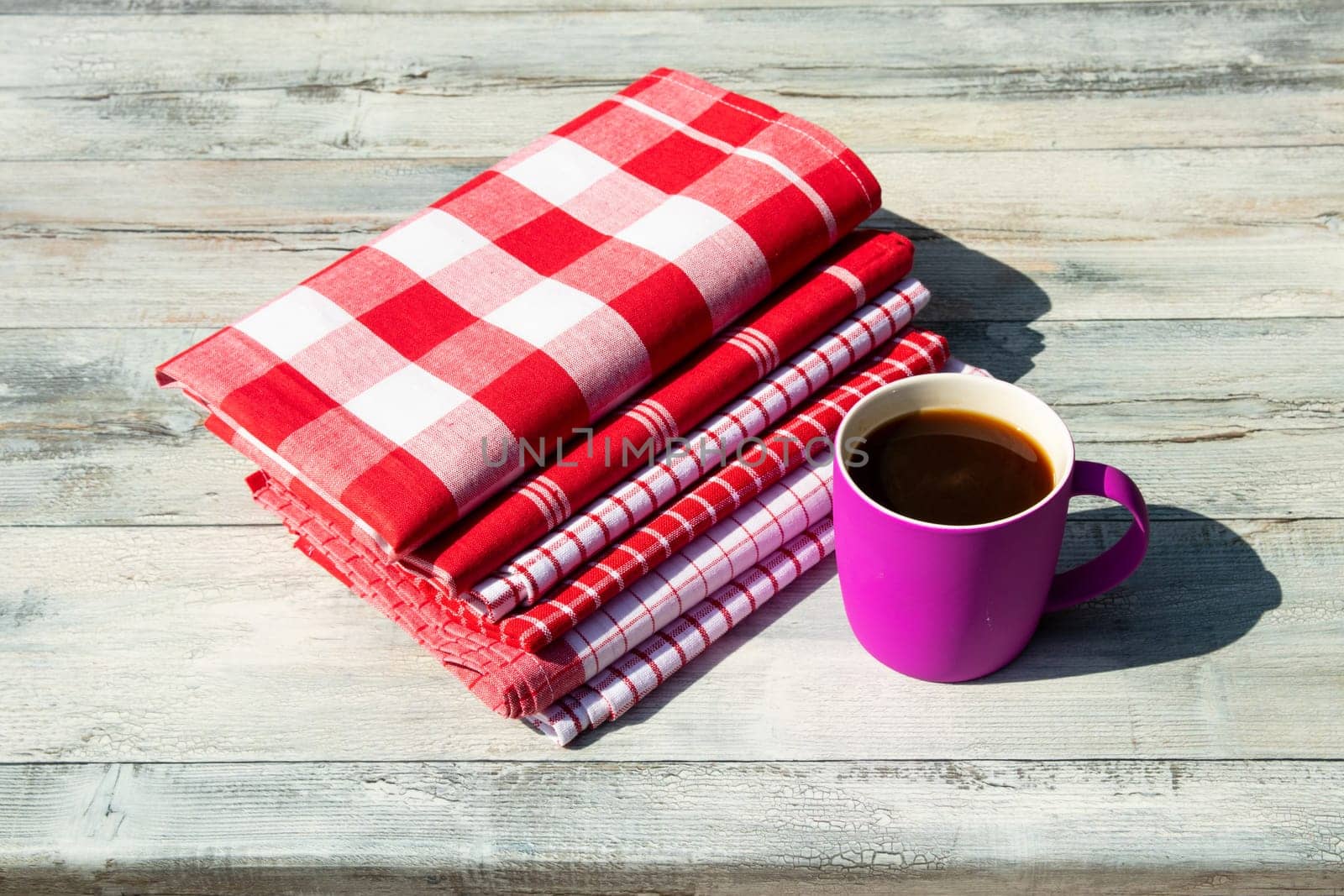 Stack of red white checkered and striped linen tableclothes and a cup of coffee on rustic bright wooden table. Outdoor.