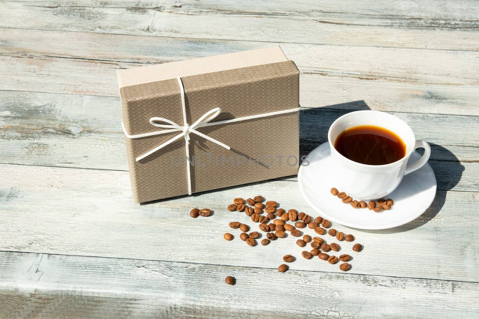 Greeting card. Birthday giftbox with white cup of black coffee with roasted beans on rustic bright wooden table. Outdoor. Card concept.