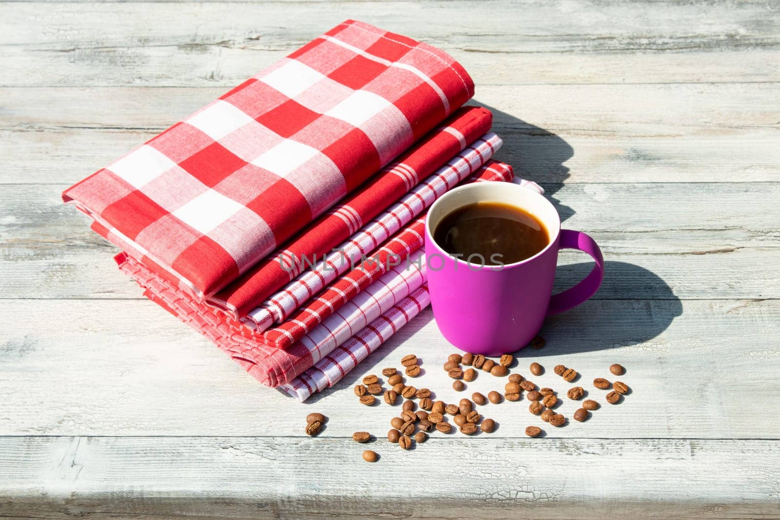 Purpure cup of hot black coffee with roasted coffee beans together with a Stack of red white checkered and striped linen tableclothes on rustic bright wooden table. Outdoor