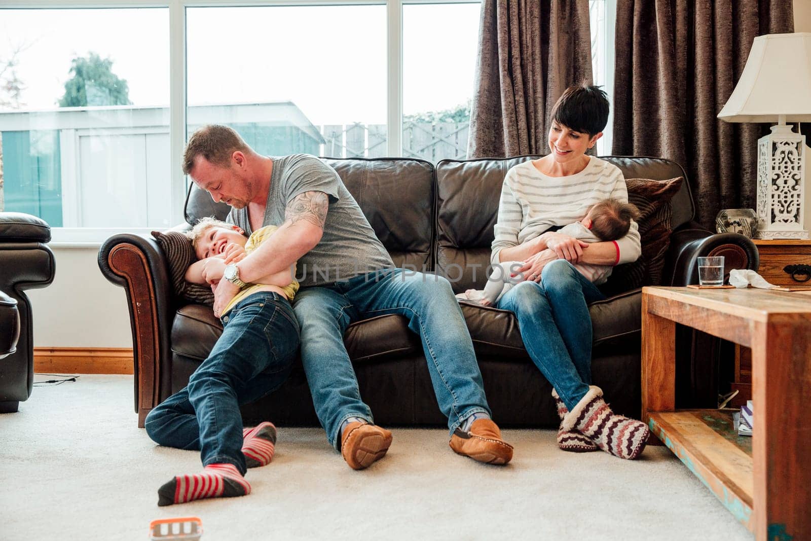Couple are relaxing at home with their children. The mother is breastfeeding the baby while watching her husband and son playfight.