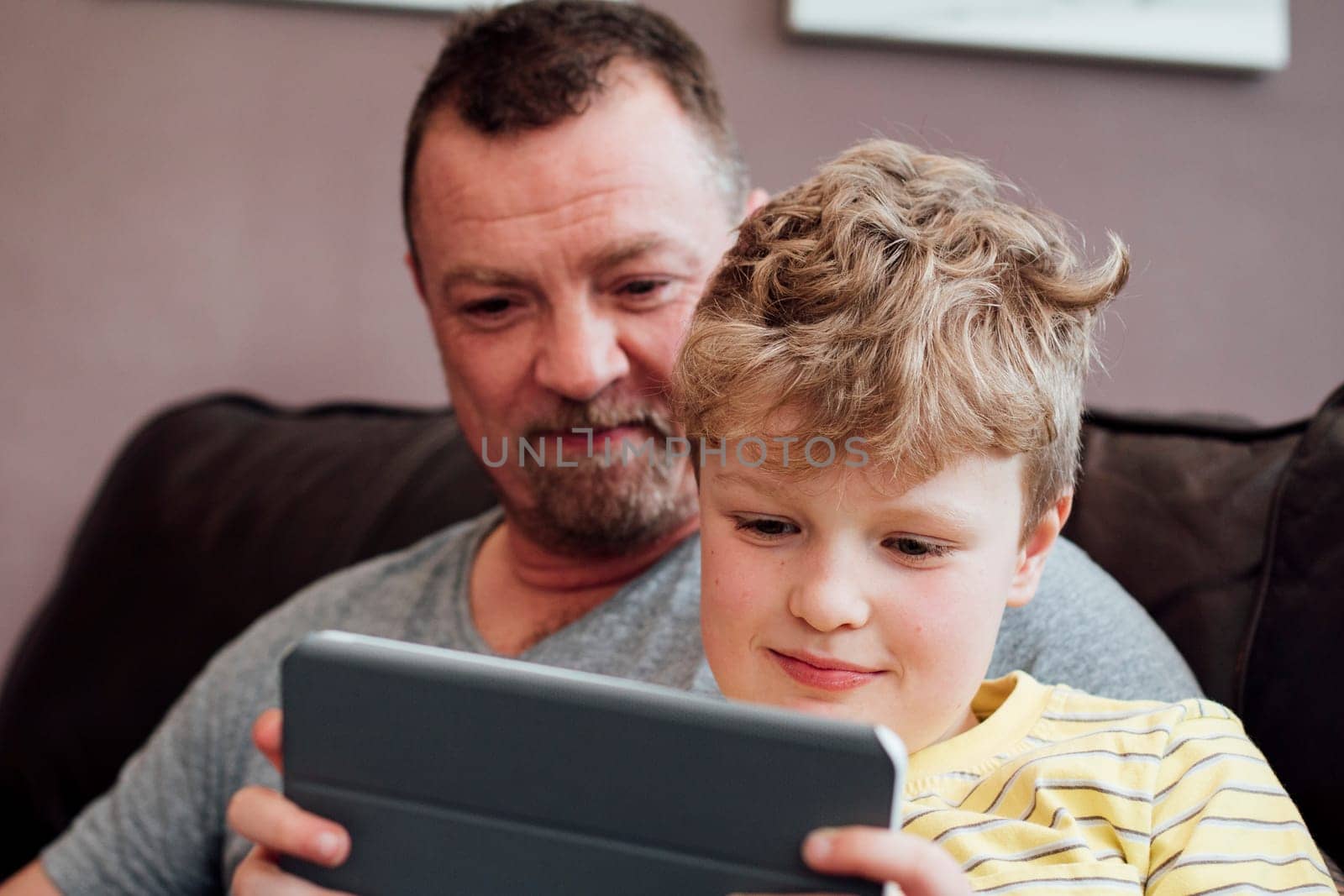 Little boy is sitting on his dads knee on the sofa at home, using a digital tablet. His dad is watching him.